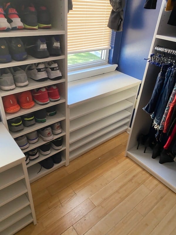 Organized walk-in closet with white shelving units displaying sneakers and folded clothes, built-in drawers under a window, and hanging garments on the side.