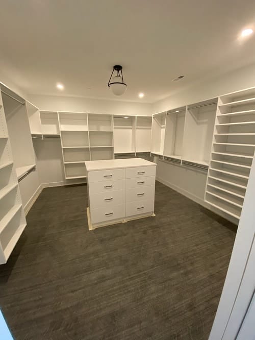 Spacious walk-in closet with white built-in shelving and hanging rods, plus a center island with drawers under recessed ceiling lights.